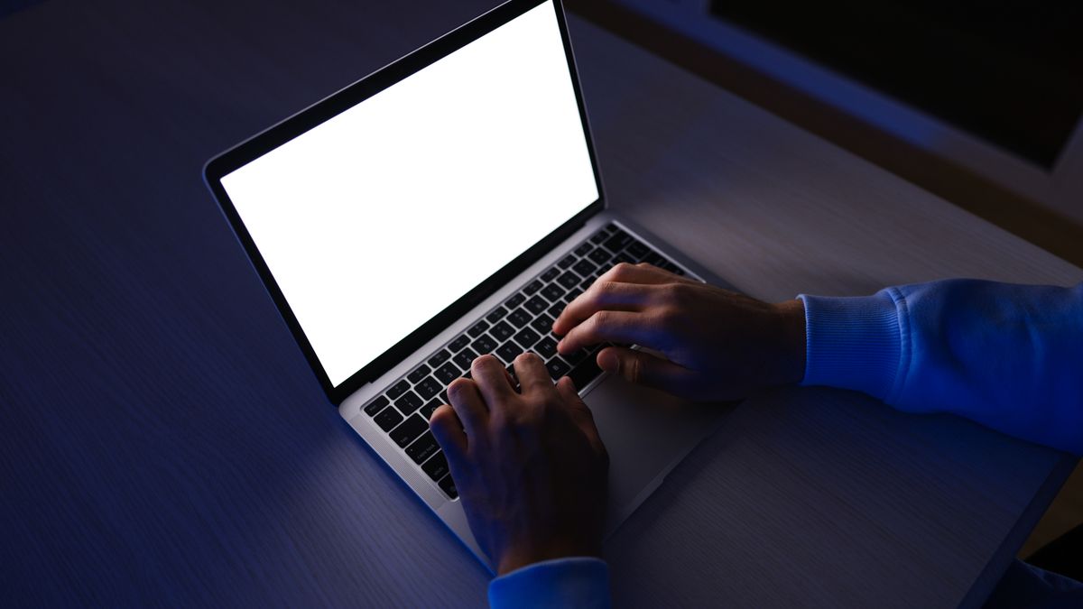 Dłonie piszące na laptopie w ciemnym pokoju, ekran świeci na biało